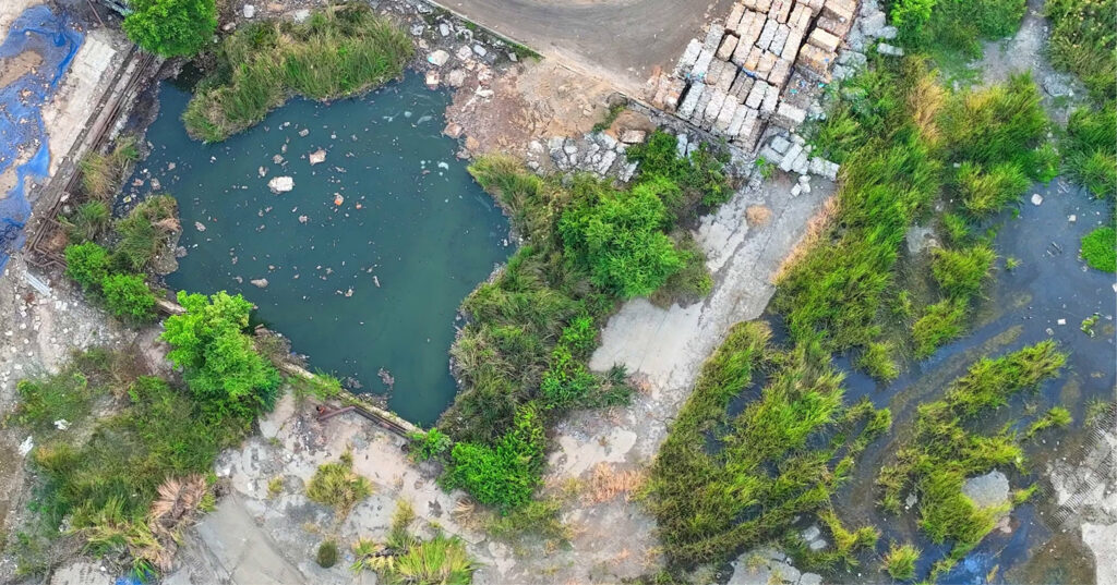 Vue aérienne d'un site pollué montrant un étang contaminé entouré de végétation et de sols dégradés, témoignant de la présence de métaux lourds issus d'activités industrielles.